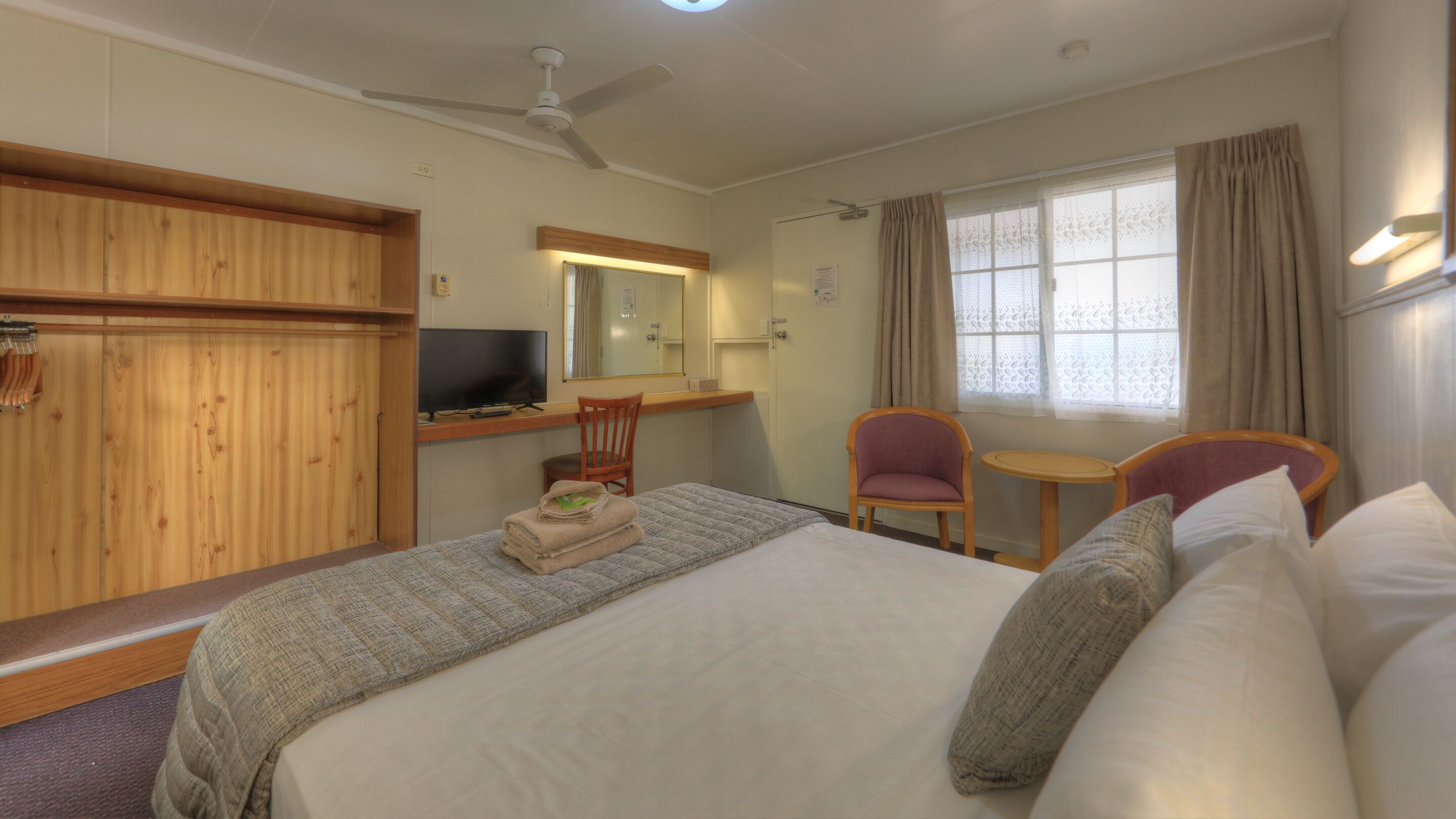 A hotel room with a bed, two chairs, a TV, a mirror, and a window with curtains.