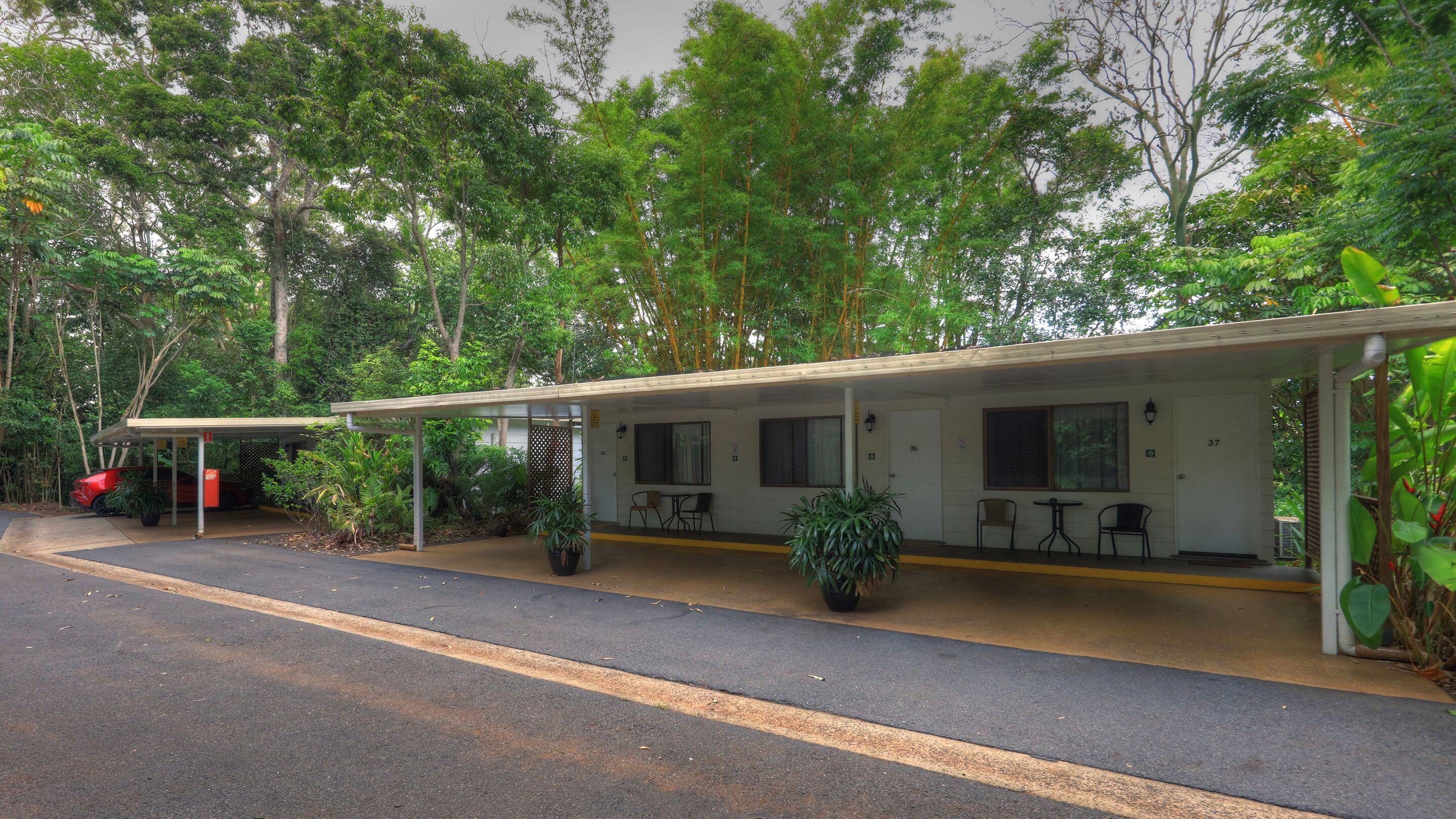 A motel with a covered outdoor seating area, two rooms, and a red car parked outside.