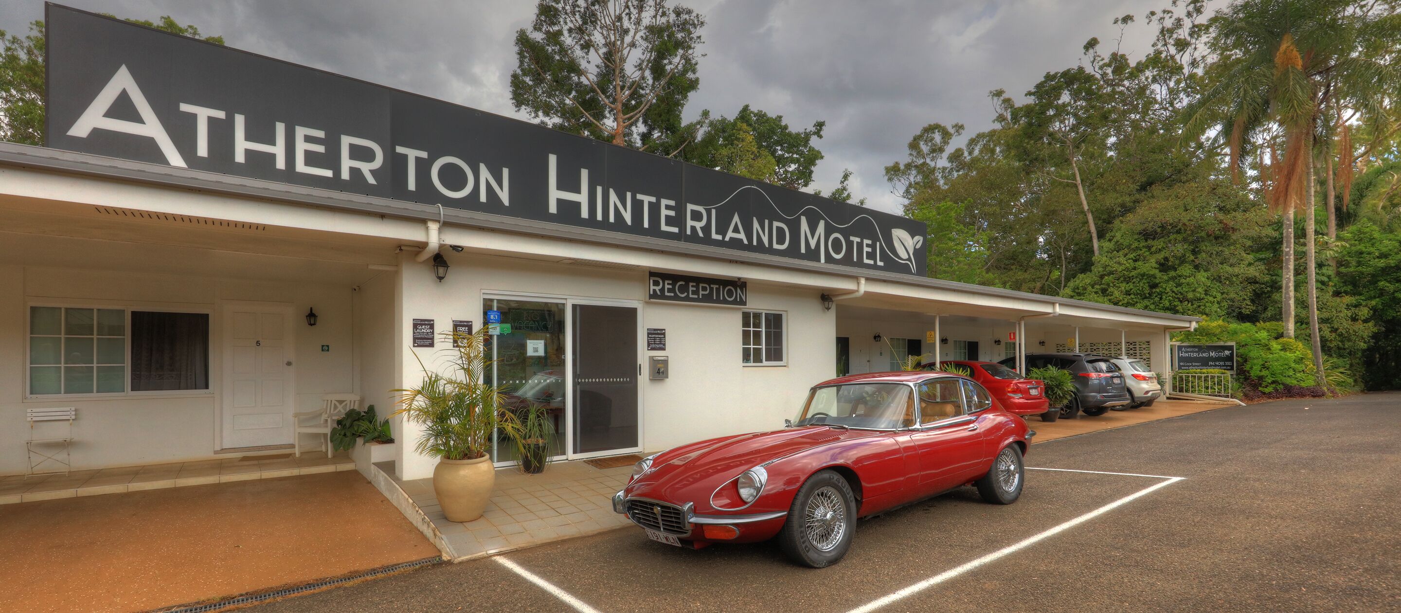 The exterior of the Atherton Hinterland Motel with a reception area, a red classic car parked in front, and several cars in the parking lot.