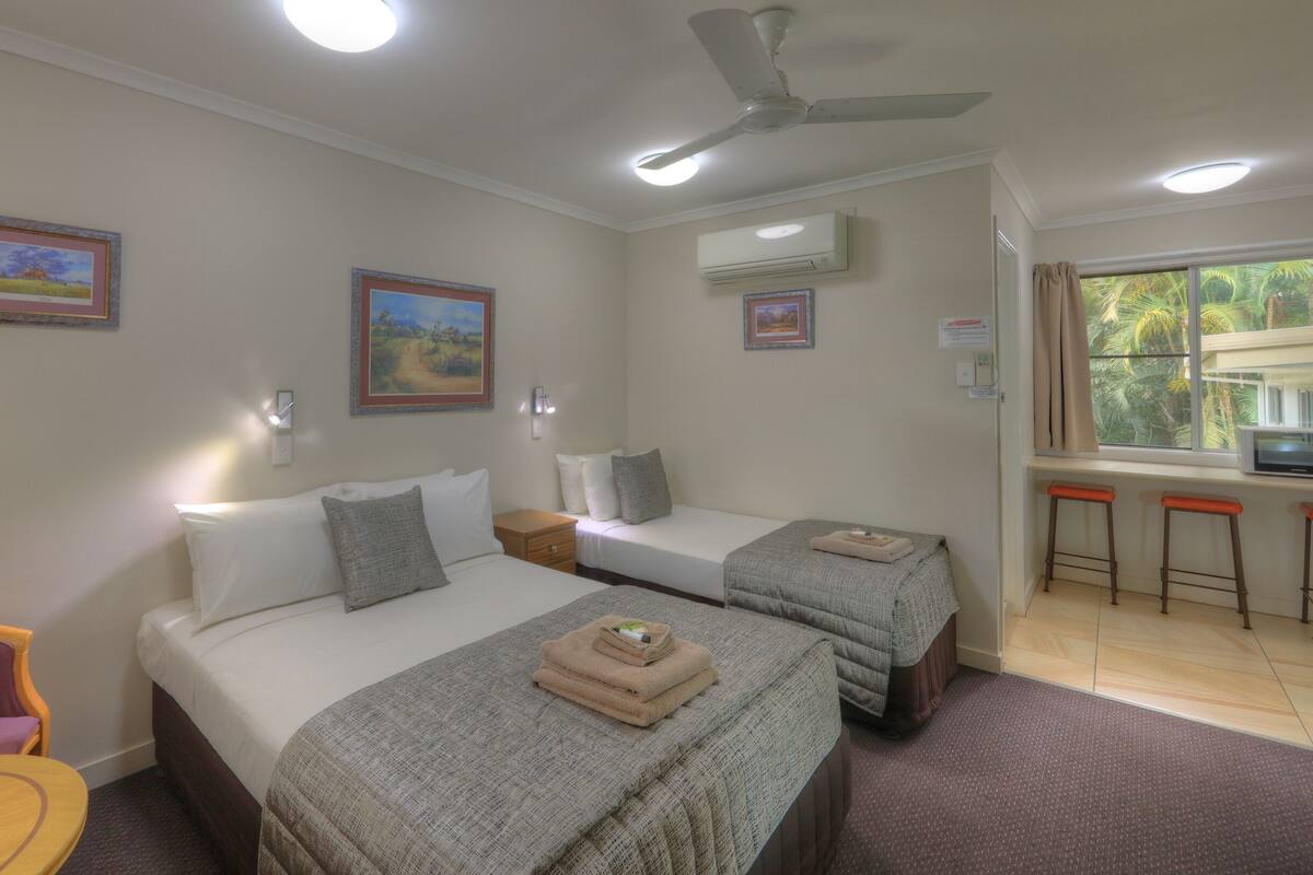 Habitación de hotel con dos camas, ventilador de techo, aire acondicionado, ventana con vistas a palmeras y una pequeña zona de kitchenette.