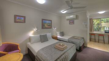 A hotel room with two beds, a ceiling fan, an air conditioning unit, a window with a view of palm trees, and a small kitchenette area.