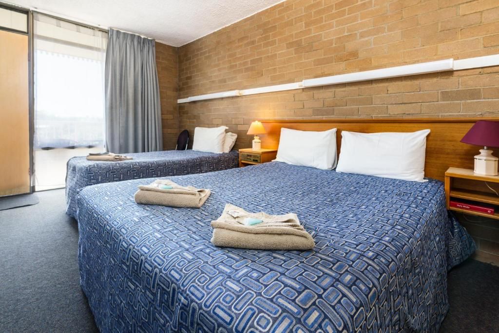 A hotel room with two beds, a brick wall, a window with curtains, and a nightstand with a lamp.