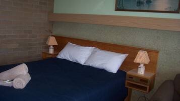 Habitación de hotel con una cama, dos almohadas, una mesita de noche con una lámpara y un cuadro en la pared.