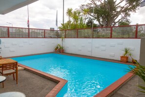 Ein rechteckiger Pool mit einer gefliesten Kante, umgeben von einer weißen Mauer mit rotem Zierstreifen. Auf der Mauer stehen Kübelpflanzen und links eine Holztischgruppe.