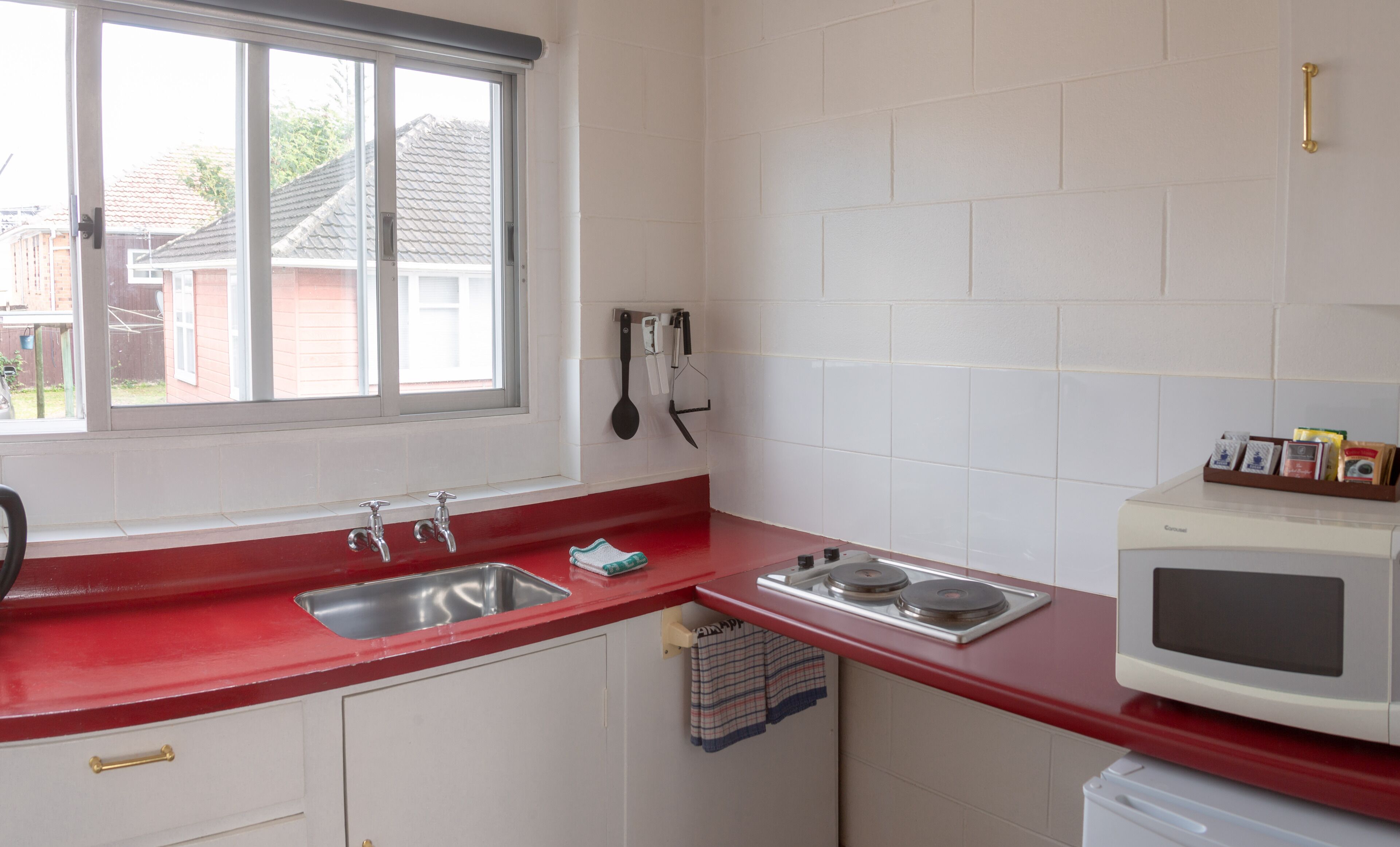 A kitchen with a red countertop, white cabinets, a stainless steel sink, a microwave, and a window with a view of a house and trees.