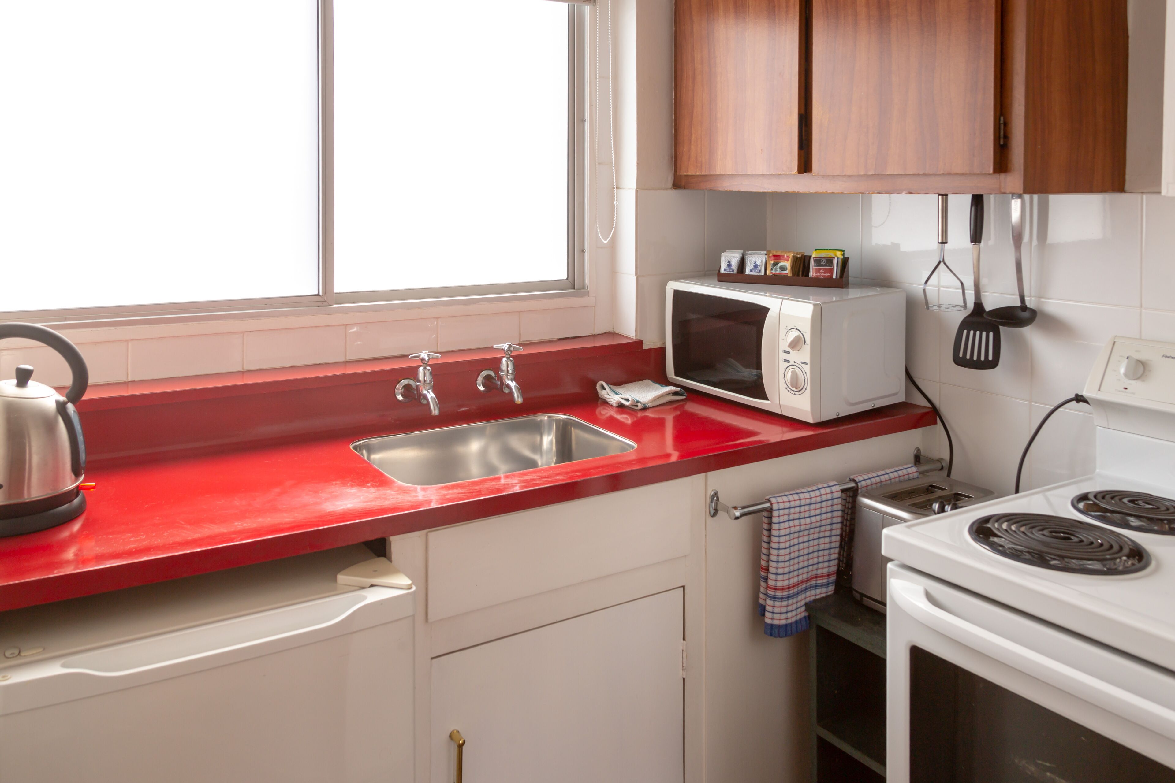 Una cocina con encimera roja, pileta de acero inoxidable, electrodomésticos blancos y muebles de madera.