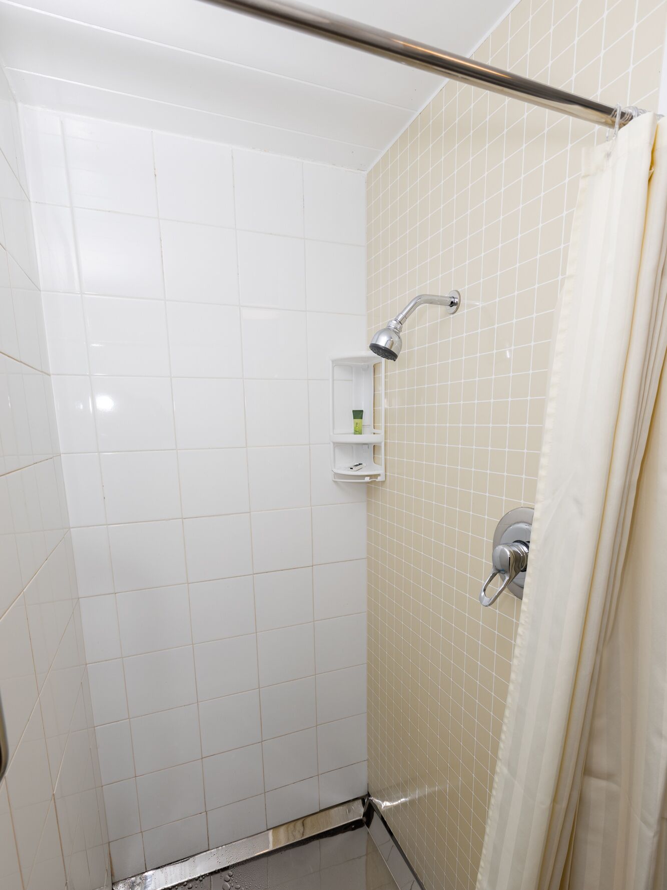 A small, tiled shower area with a handheld showerhead, a white shelf with toiletries, and a shower curtain.