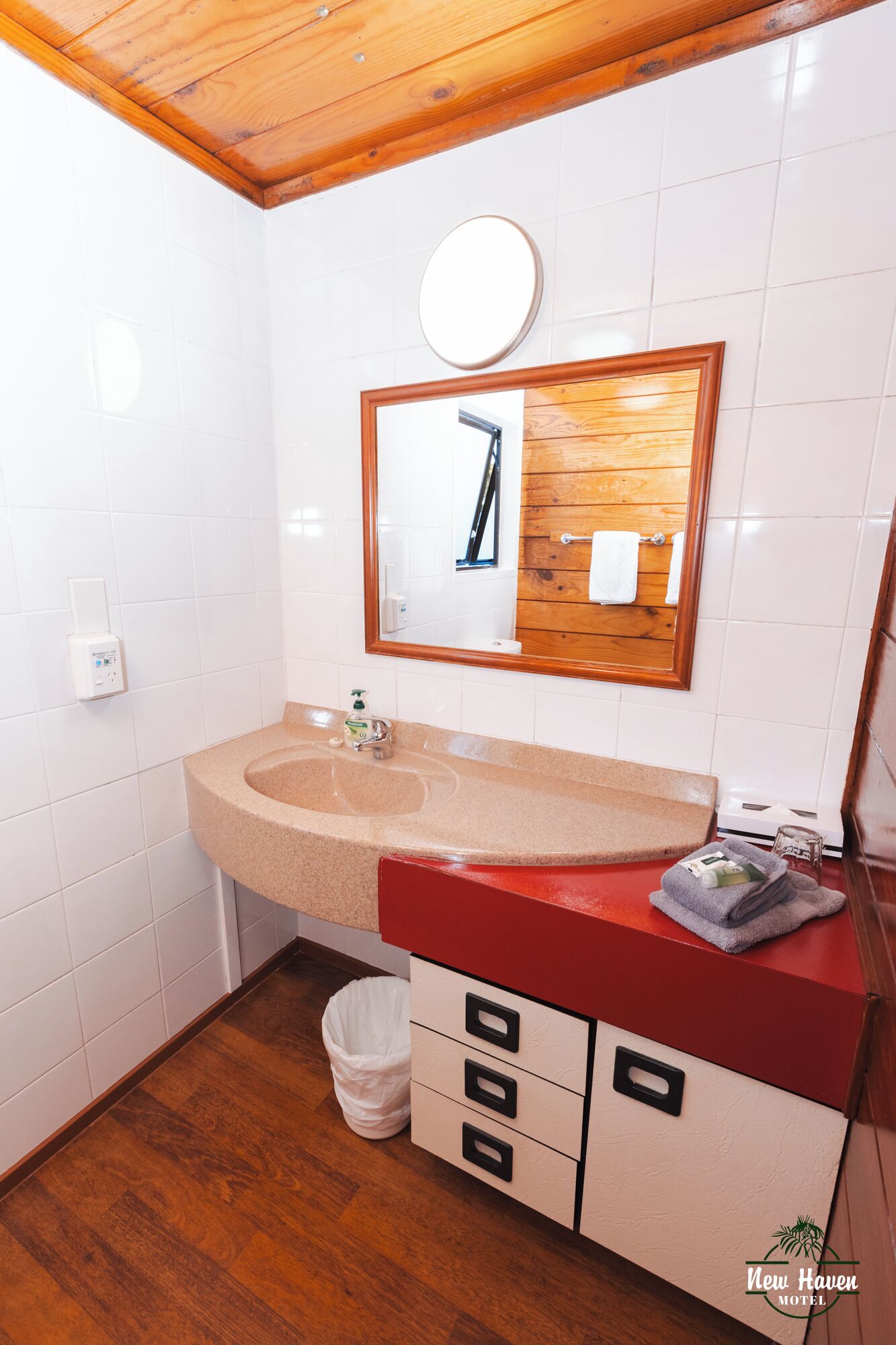 A bathroom with a wooden ceiling, white tiled walls, a beige sink, a red countertop, a mirror, and a trash bin.