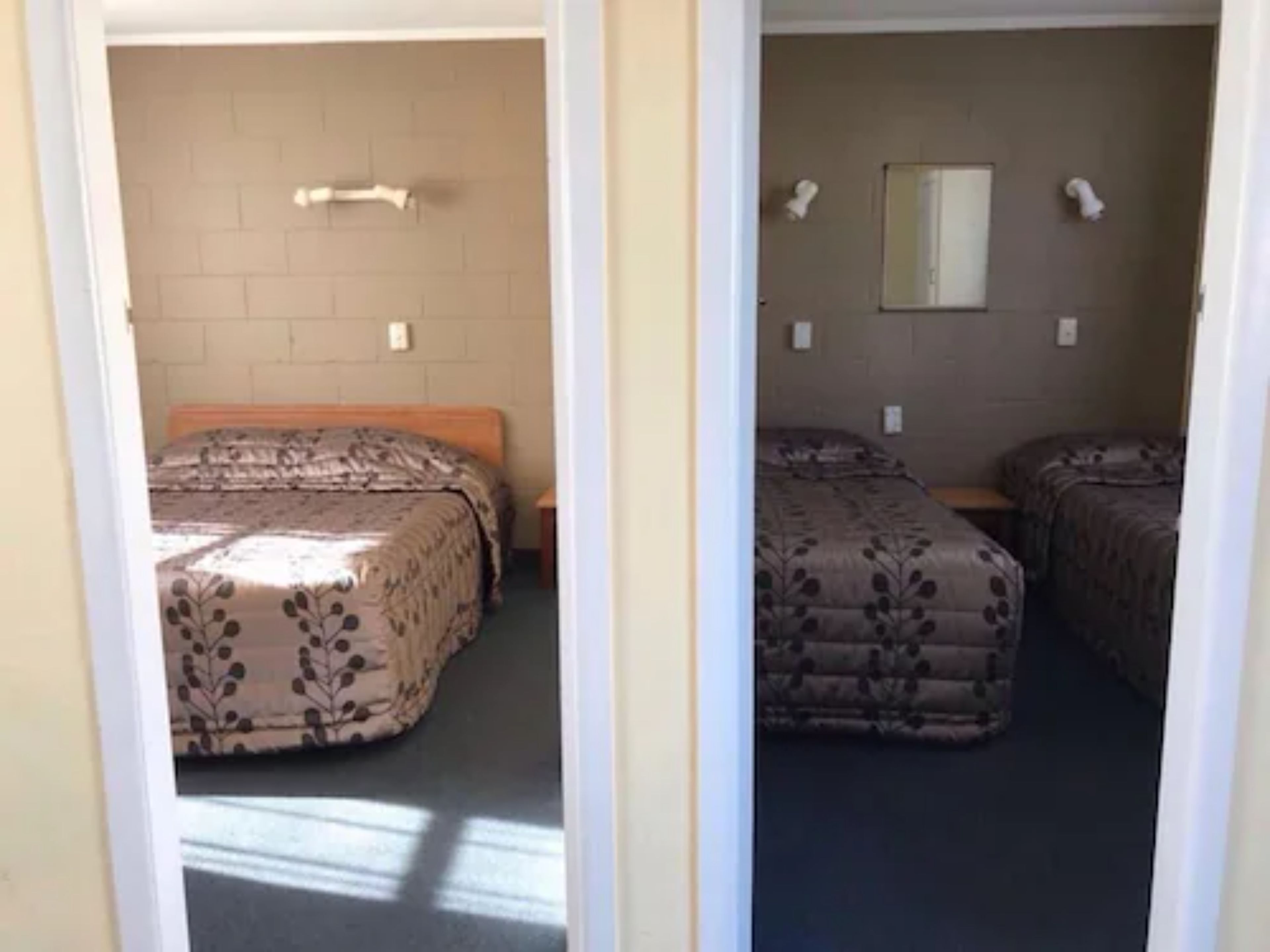 Une chambre avec deux lits, chacun recouvert d’une couvre-lit à motifs, une petite table et un luminaire fixé au mur.