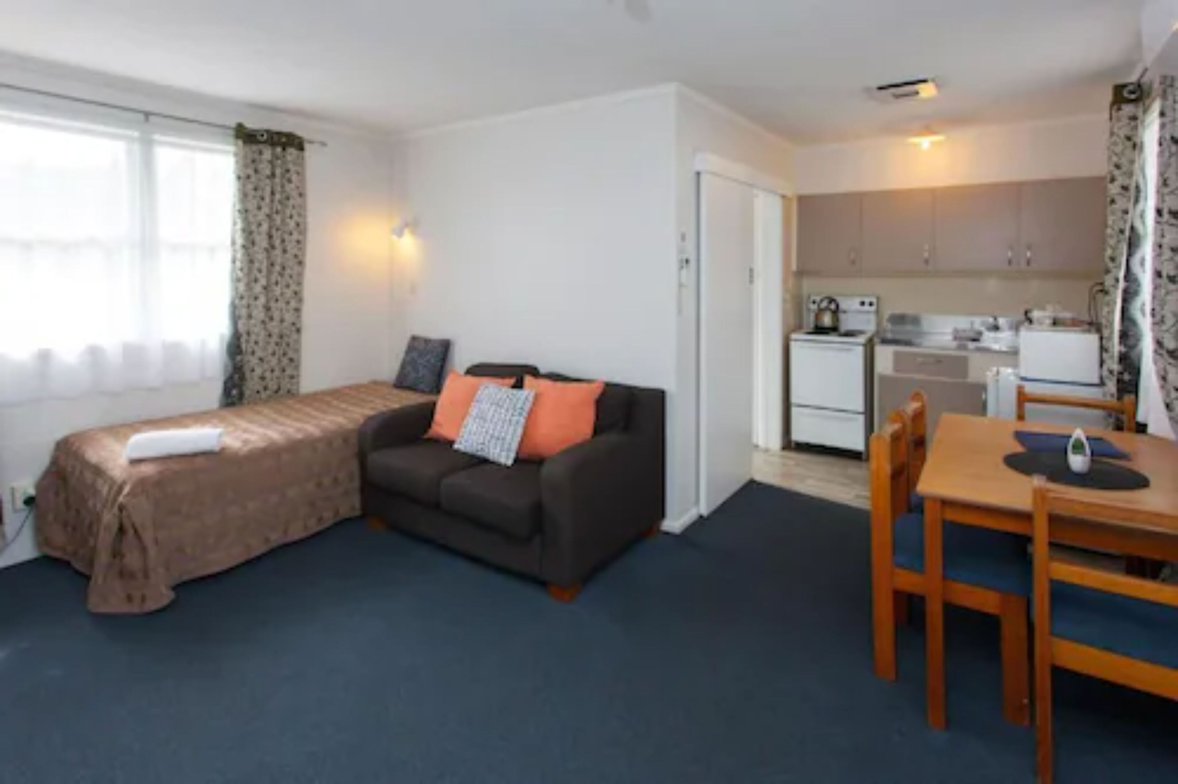 A compact room with a bed, a sofa, a dining table with chairs, a kitchenette, and a window with curtains.