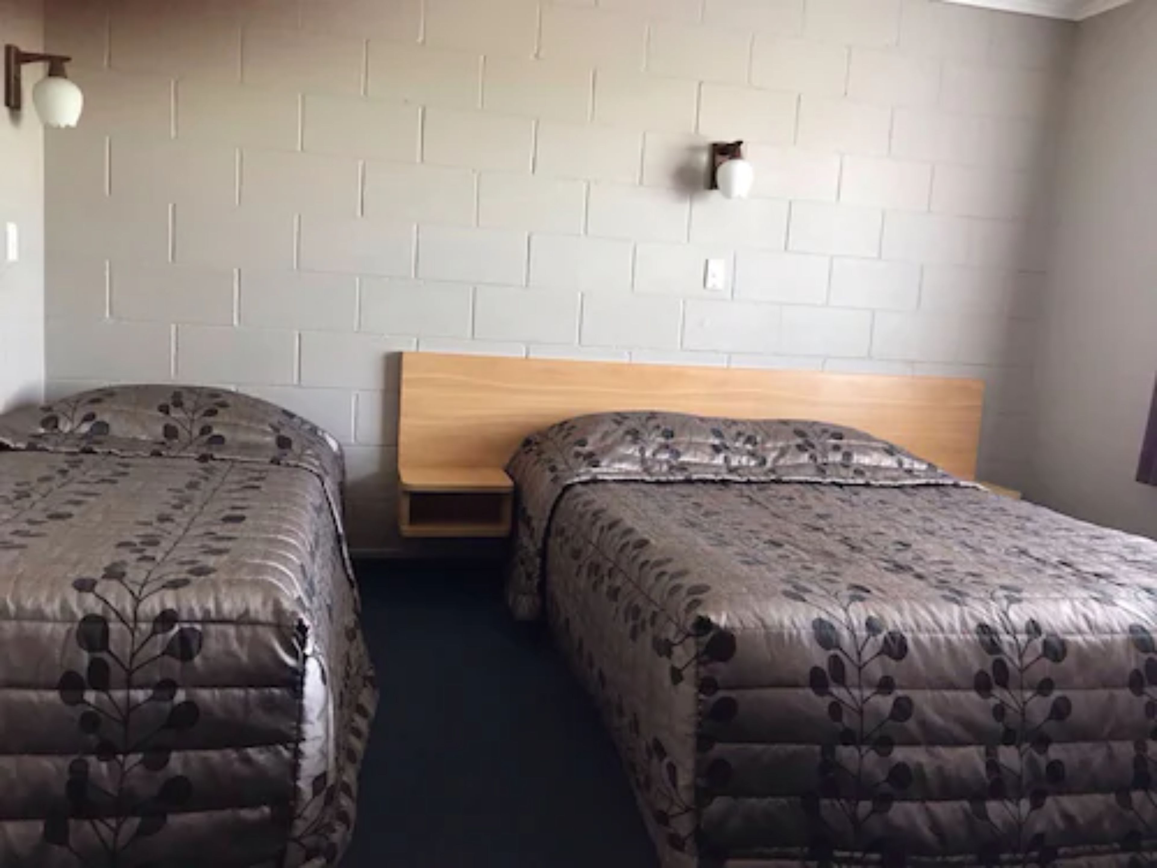 A room with two beds, each with a wooden headboard and floral-patterned bedding. There are wall-mounted light fixtures and a small wooden shelf between the beds.