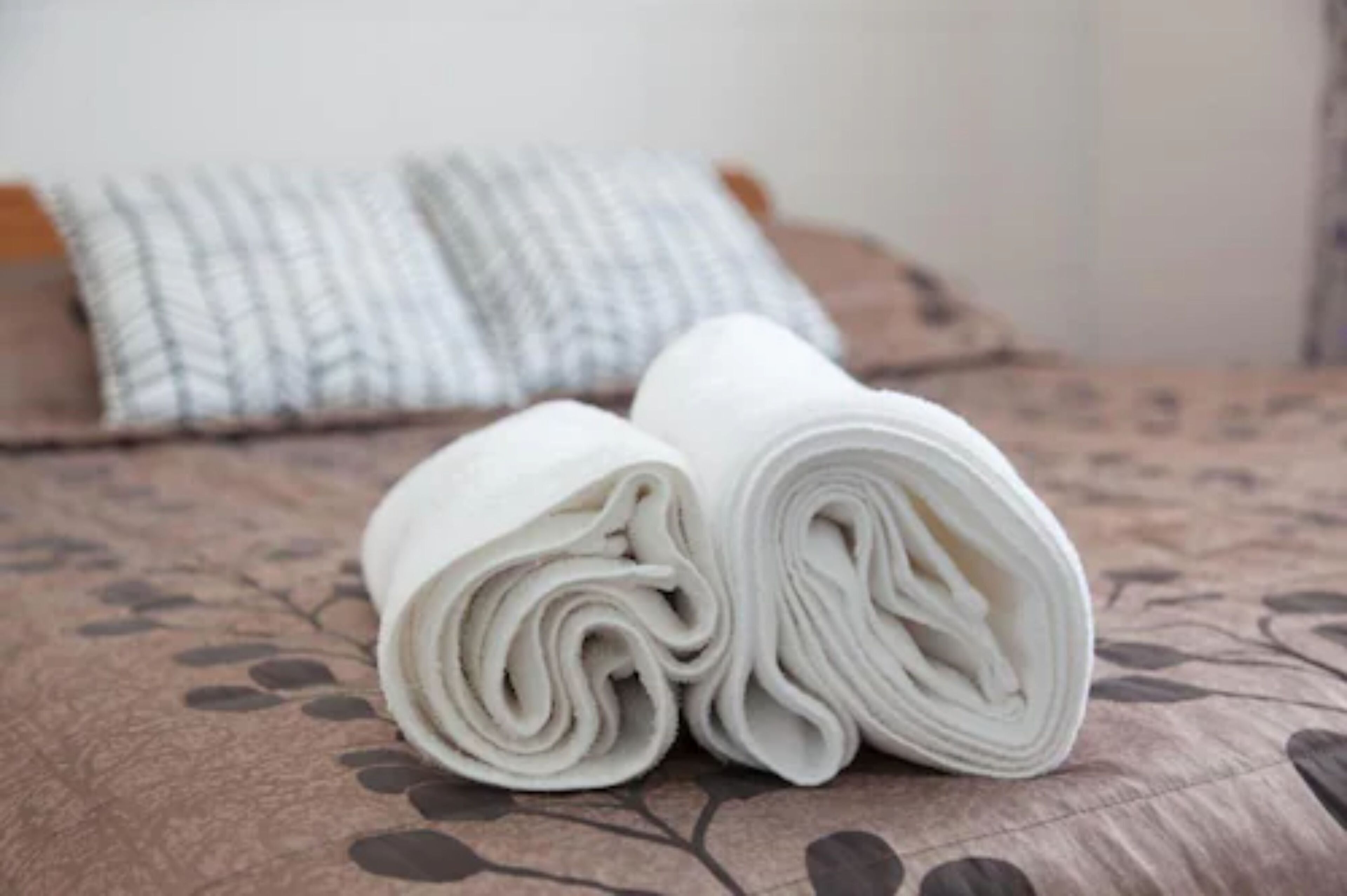 Una cama con una toalla blanca enrollada y un cojín estampado sobre una colcha con motivos florales.
