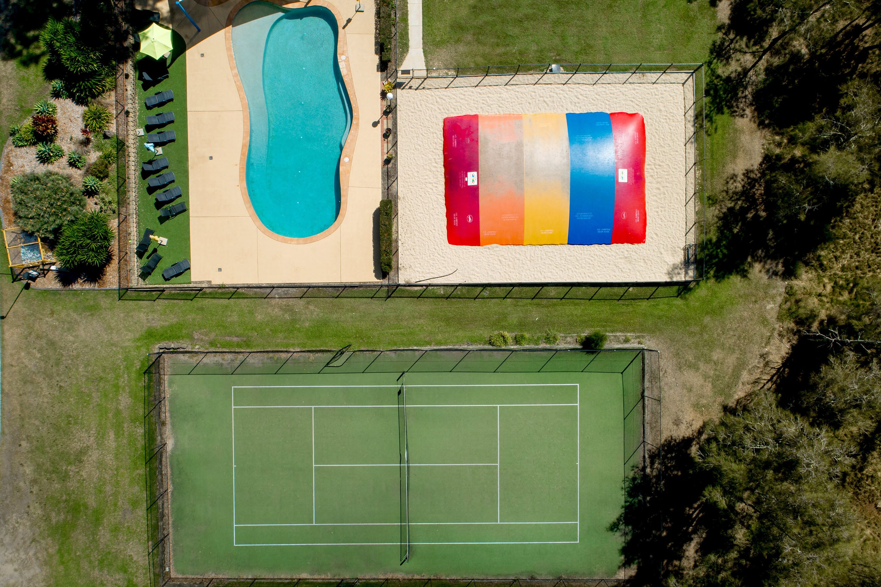 An aerial view of a property with a swimming pool, a tennis court, and a multi-colored sand volleyball court.