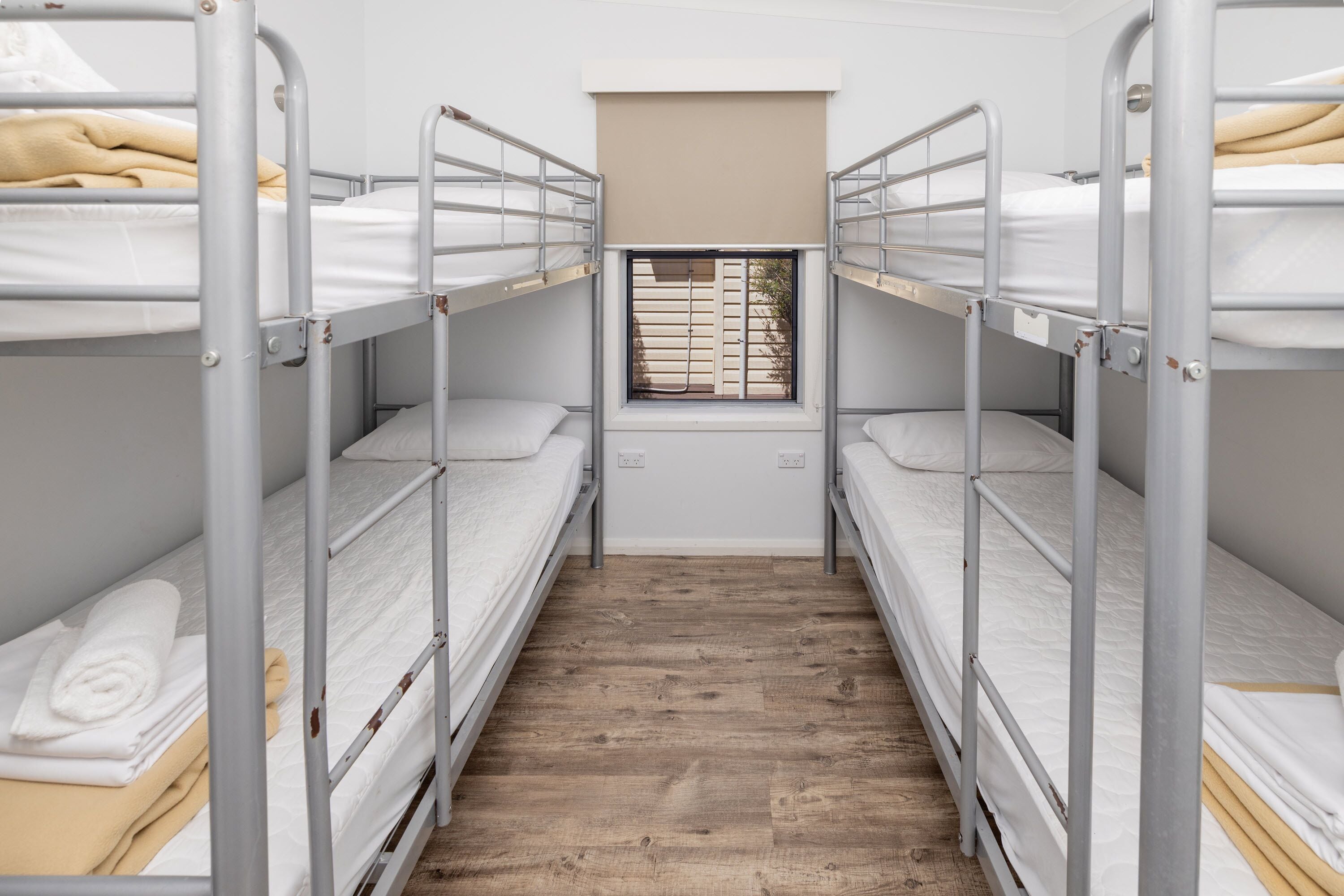 A dormitory room with bunk beds, a window with blinds, and a wooden floor.