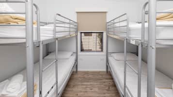 A dormitory room with bunk beds, a window with blinds, and a wooden floor.