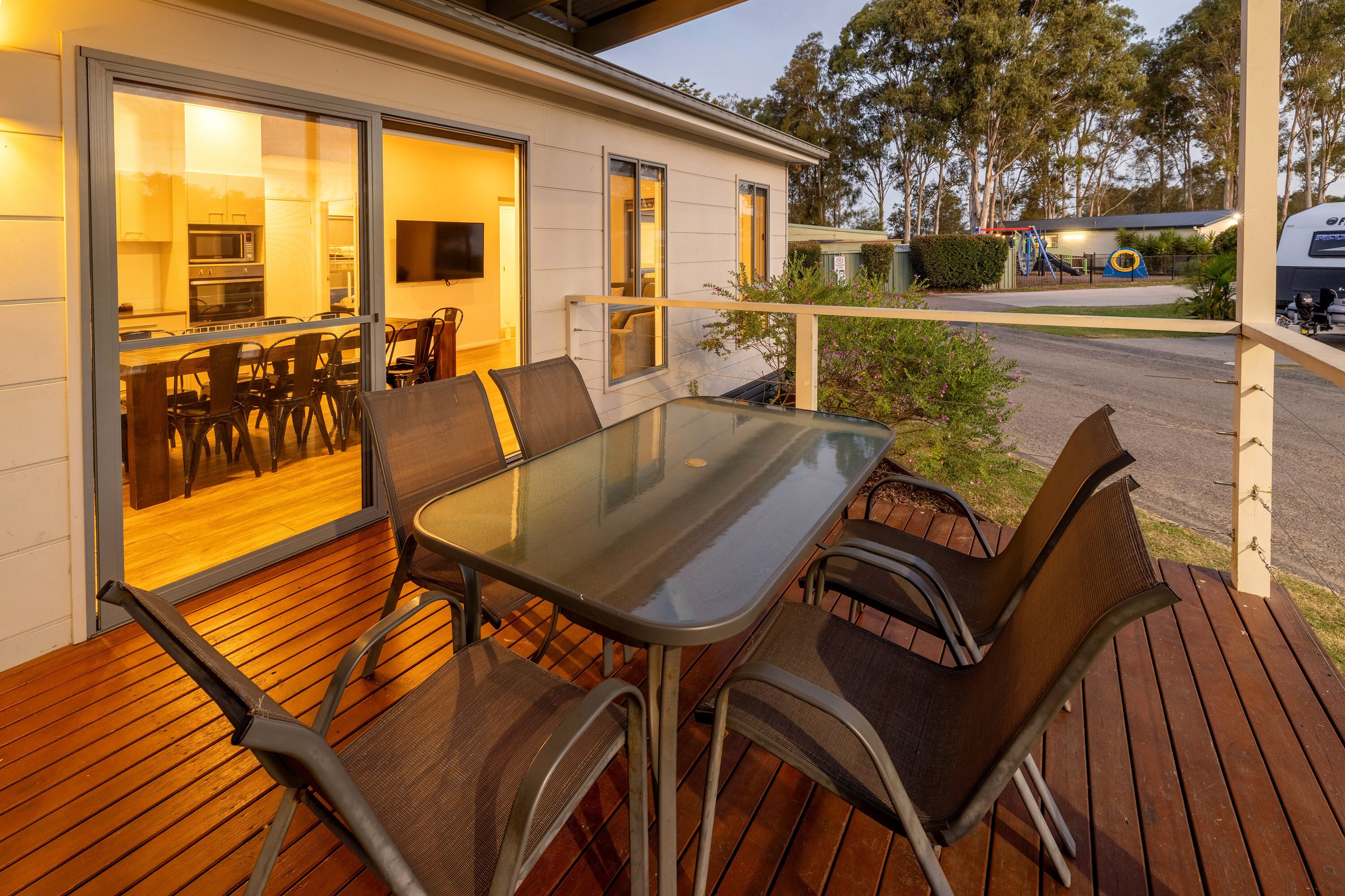 A wooden deck with a glass table and chairs, a sliding glass door leading to a kitchen, and a playground in the background.