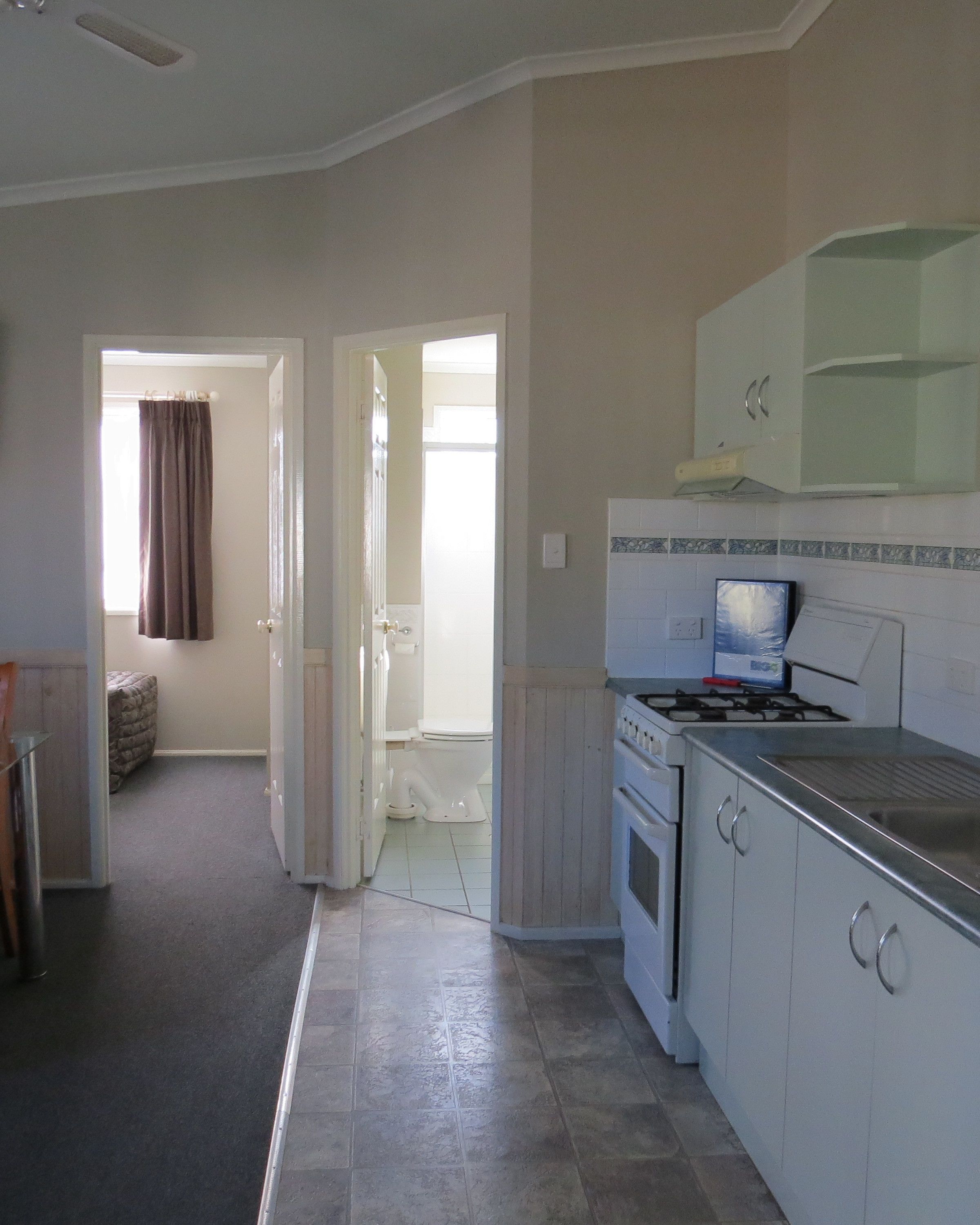 A compact kitchen with white cabinets, a stove, and a microwave. There is a doorway leading to a bathroom with a toilet and a shower.
