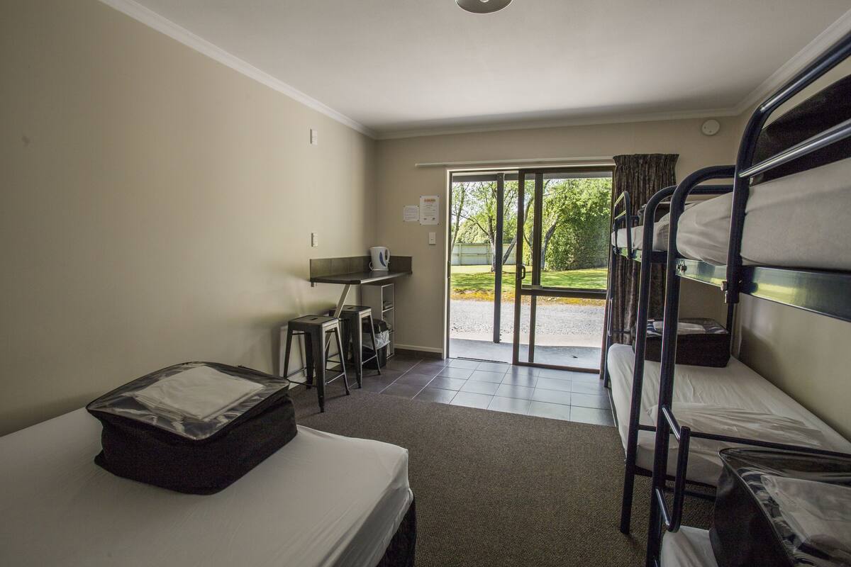 A dormitory room with bunk beds, a desk, a chair, and a window with a view of a garden.