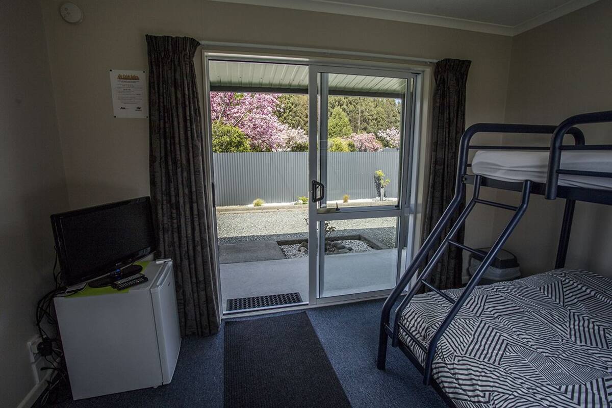 A room with a bunk bed, a small desk with a television and remote control, and a sliding glass door leading to an outdoor area with a view of trees and a fence.
