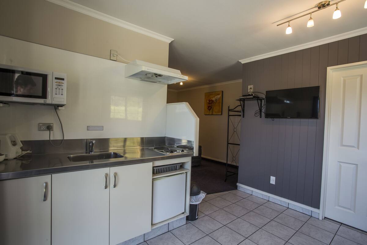 A compact kitchen with a microwave, sink, and stove. There is a TV mounted on the wall and a door to the right.