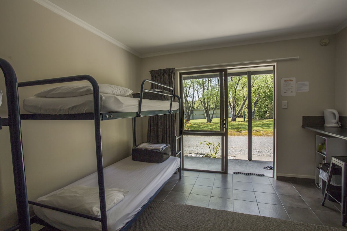 A dormitory room with bunk beds, a sliding glass door leading to an outdoor area, a small kitchenette, and a bulletin board.