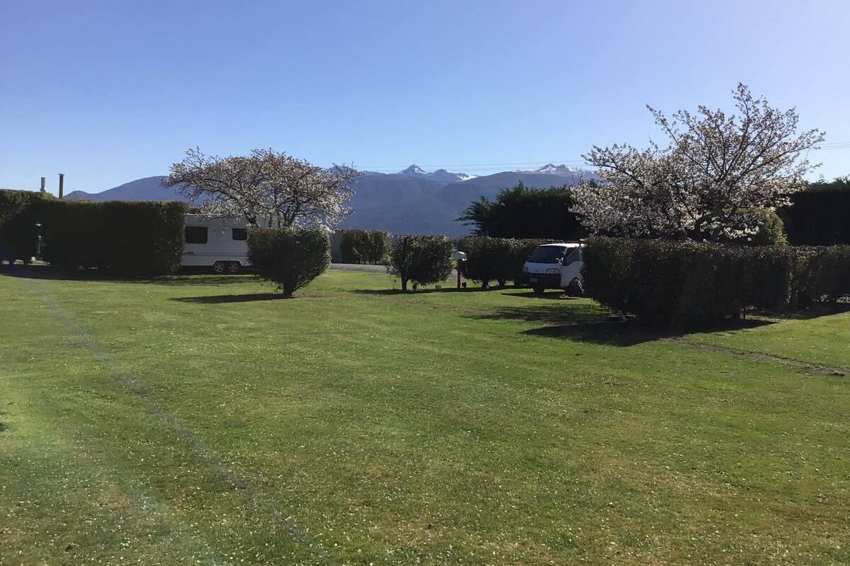 A grassy field with a white RV and a white car parked among trees and a mountain range in the background.