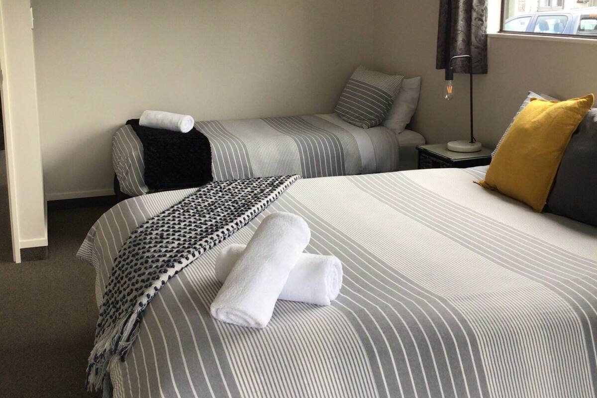 A hotel room with two beds, a painting on the wall, a window with curtains, and a lamp on a bedside table.
