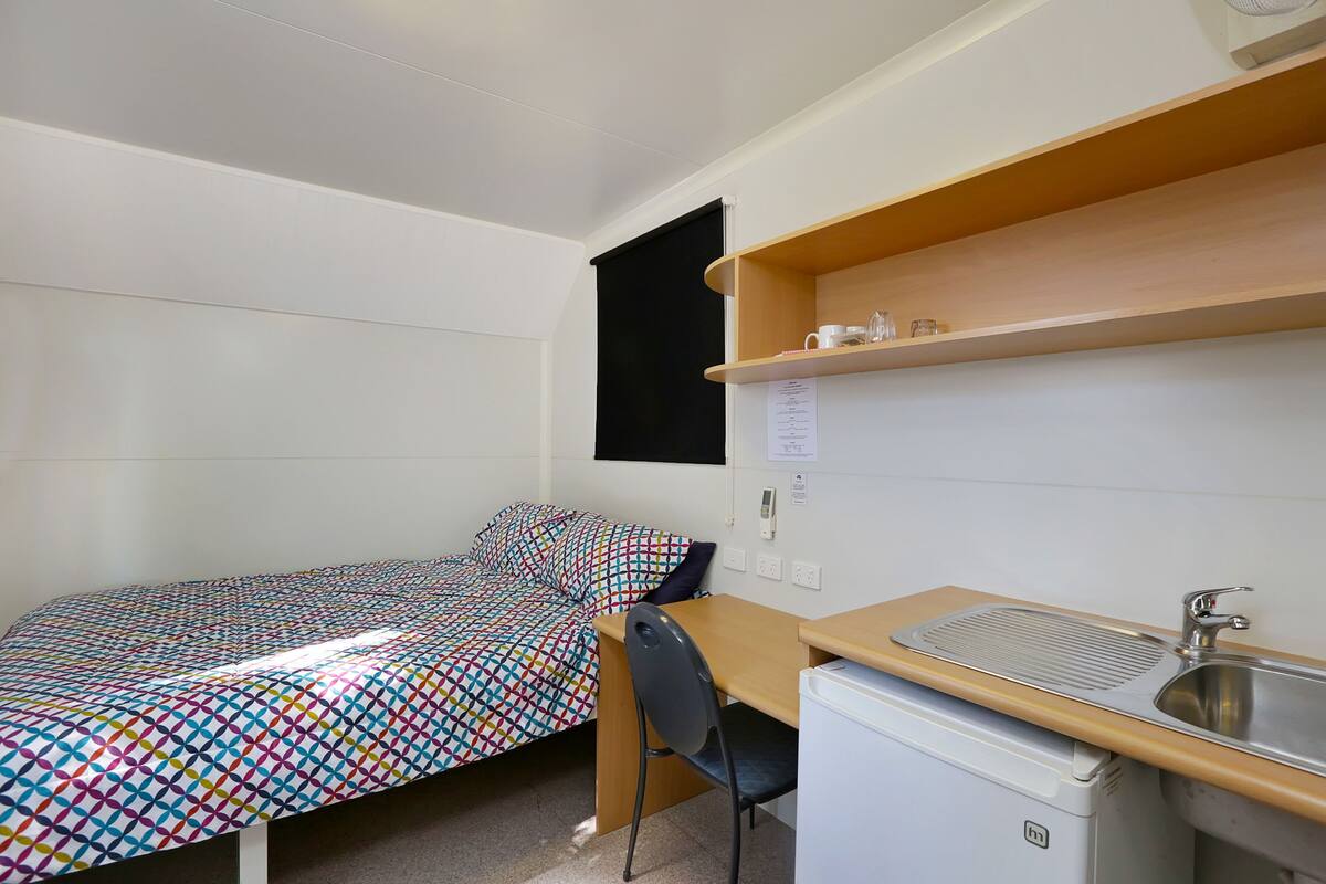 Ein kleiner, kompakter Raum mit Bett, Schreibtisch, Stuhl, Waschbecken und kleinem Kühlschrank.