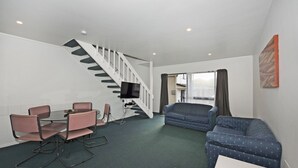 A living room with a staircase, a dining area with pink chairs, a blue sofa, and a flat-screen TV mounted on the wall. - Camellia Court Motel (Lower Hutt)