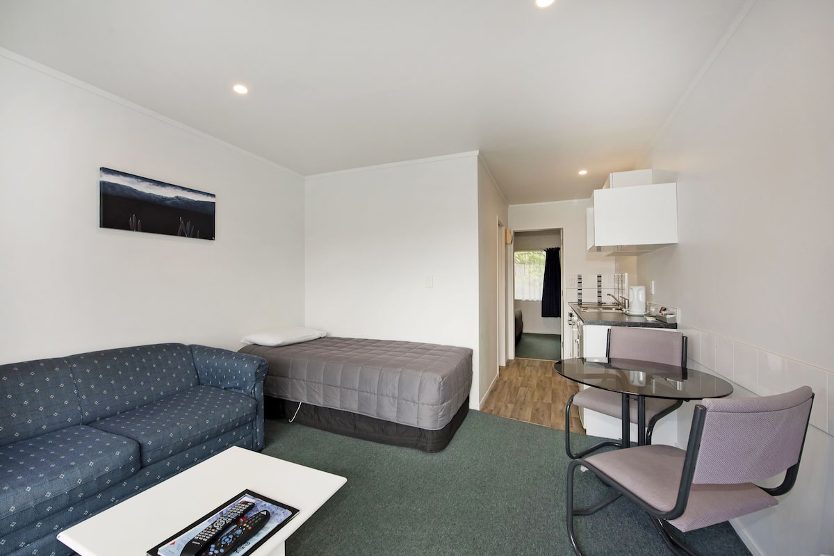 A small, modern room with a sofa, a single bed, a small table with a chair, and a kitchenette area.