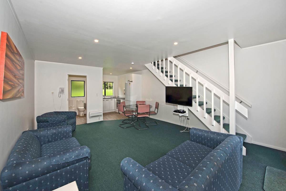 A modern living room with two blue patterned sofas, a dining table with chairs, a kitchen area, and a staircase.
