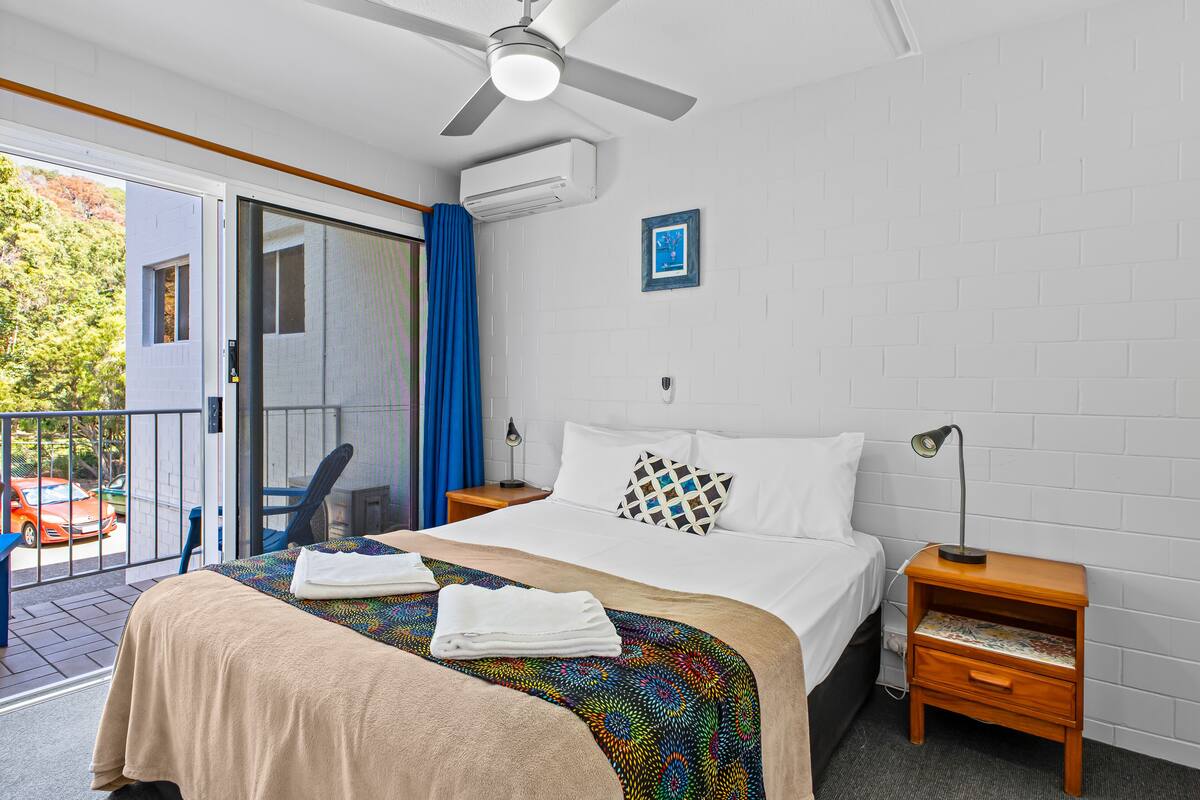 A hotel room with a bed, a nightstand, a ceiling fan, a window with curtains, and a view of the outside.
