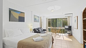 Habitación de hotel con cama, sofá, televisor, zona de comedor con mesa y sillas, y balcón con vistas a zonas verdes.
