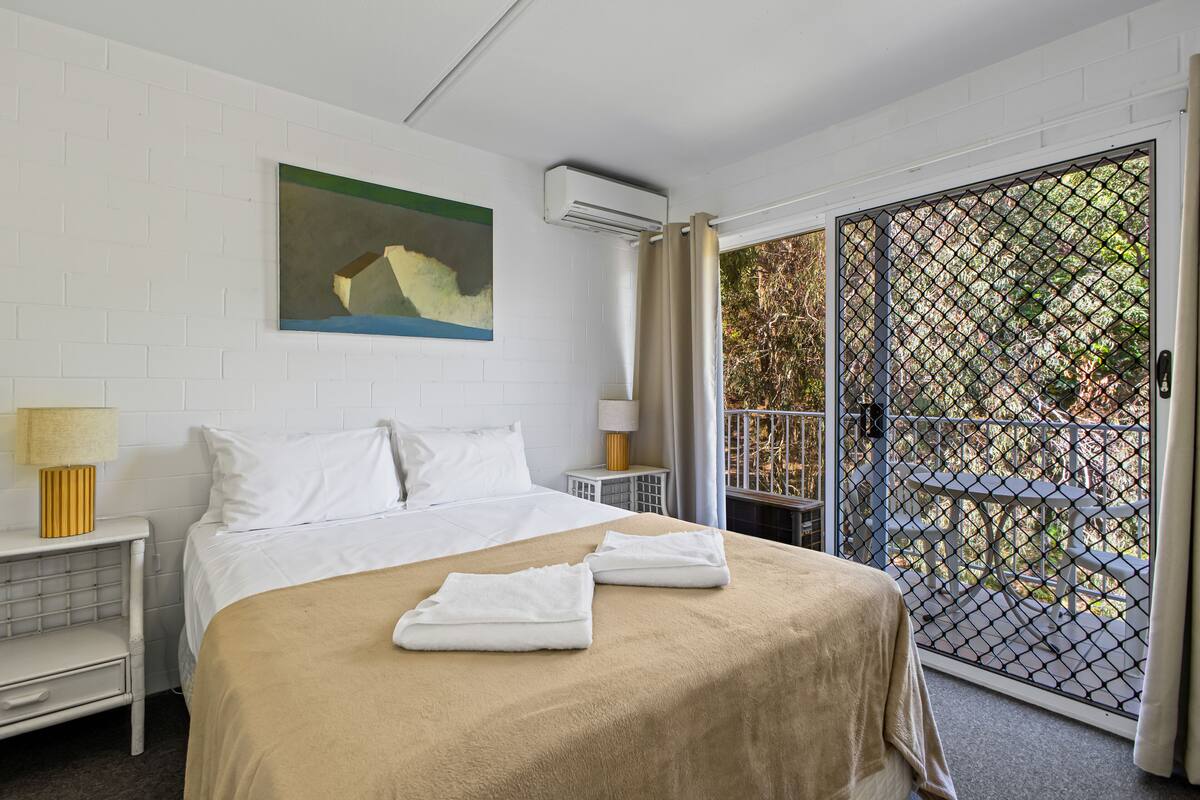 A hotel room with a bed, a painting on the wall, an air conditioning unit, a nightstand with a lamp, and a view through a glass door to an outdoor area with tables and chairs.