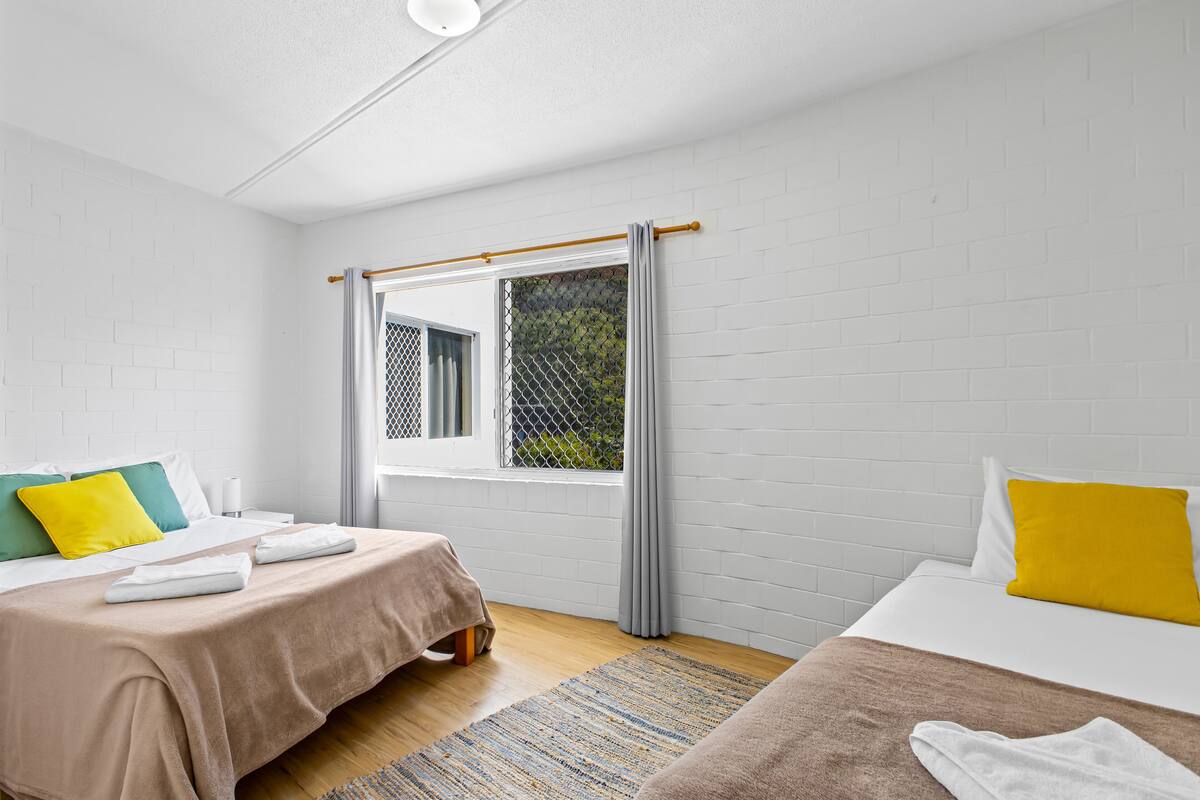 A hotel room with two beds, a window with a view of greenery, white brick walls, and a wooden floor.