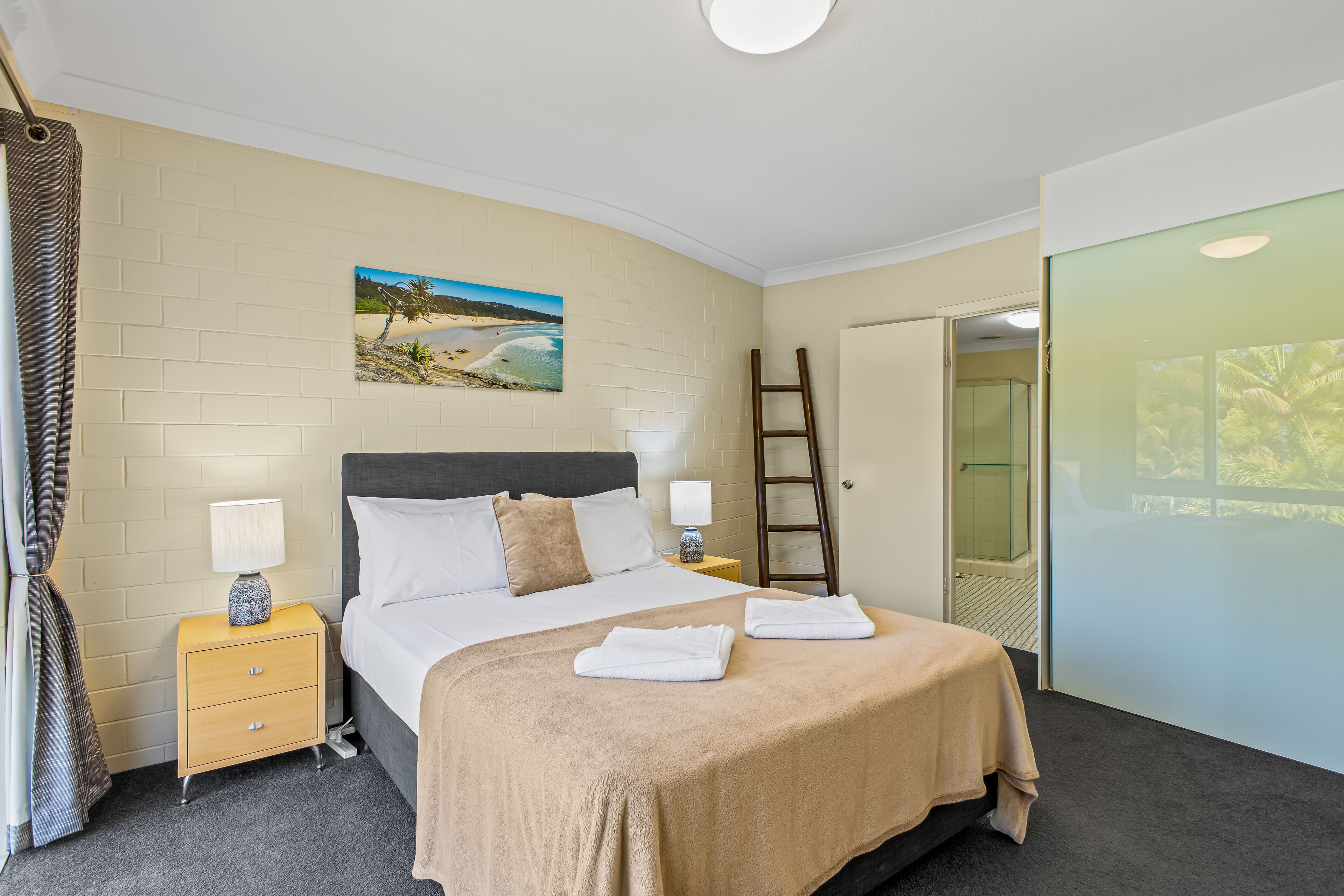 A hotel room with a bed, a nightstand, a lamp, a painting, a ladder, and a sliding door to a balcony.