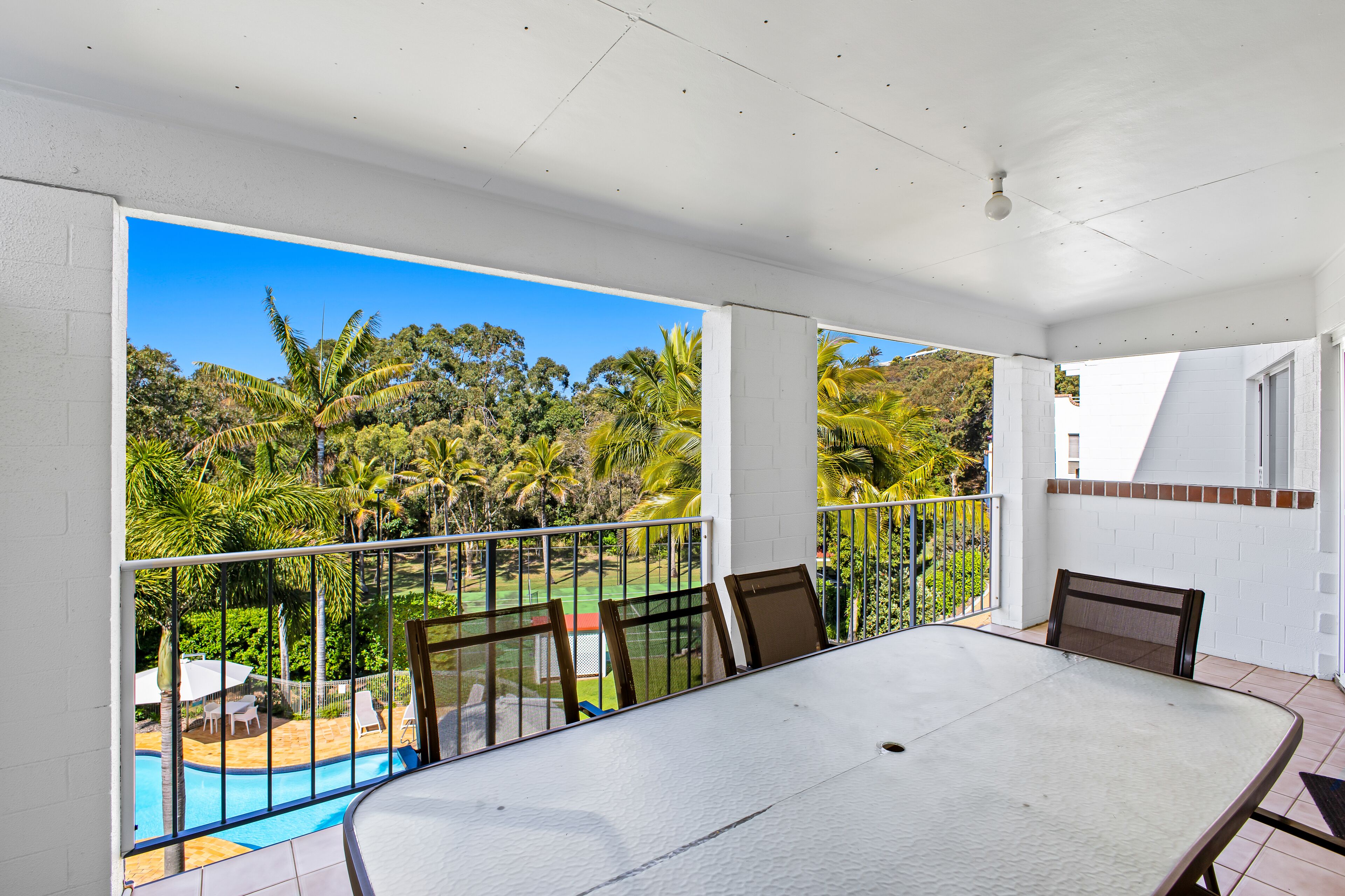 A covered outdoor area with a table and chairs, overlooking a swimming pool and lush greenery.
