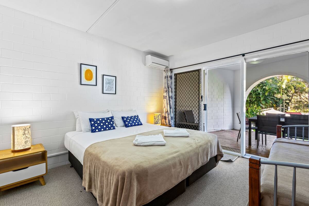 A bedroom with a bed, a nightstand, a window with a view of the outdoors, and a sliding glass door leading to an outdoor seating area.
