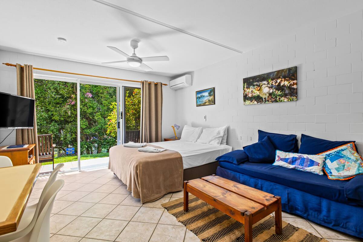 A modern hotel room with a bed, a blue sofa, a wooden table, a TV, and a large window with curtains.