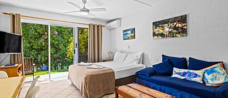 A modern hotel room with a bed, a blue sofa, a wooden table, a TV, and a large window with curtains.