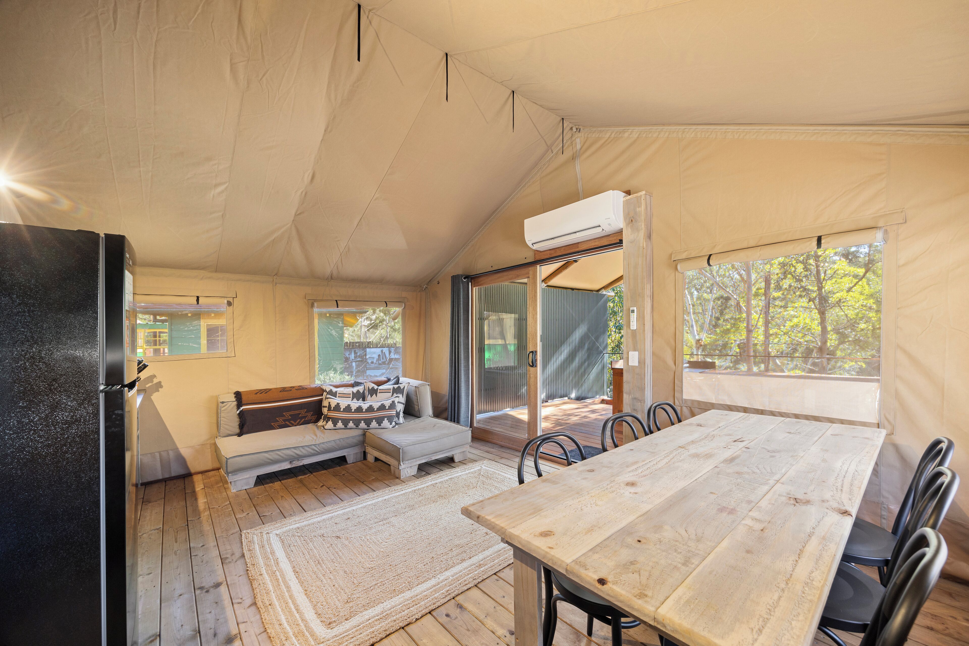 A tented accommodation with a wooden table and chairs, a sofa with cushions, and a sliding glass door leading to an outdoor area.