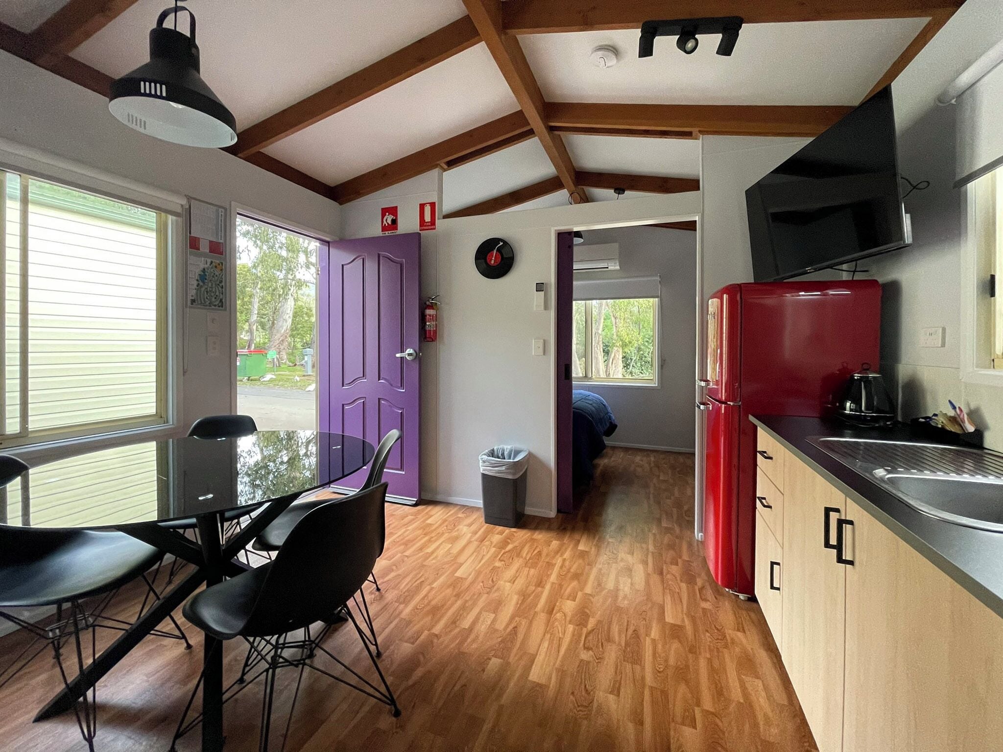 A compact kitchen with a dining area, wooden floors, a purple door, and a red refrigerator.