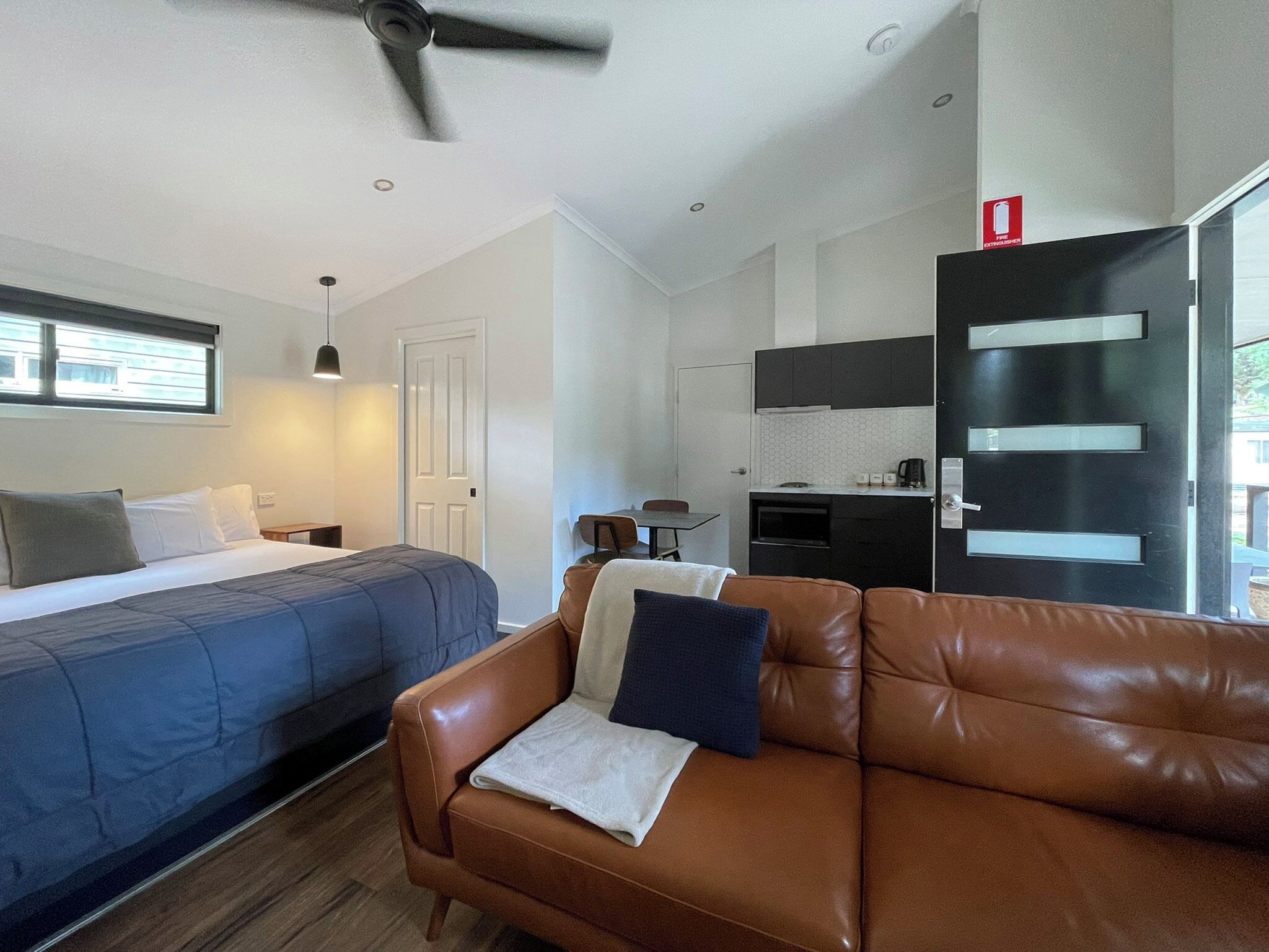 A modern hotel room with a bed, a brown leather sofa, a kitchenette, and a ceiling fan.