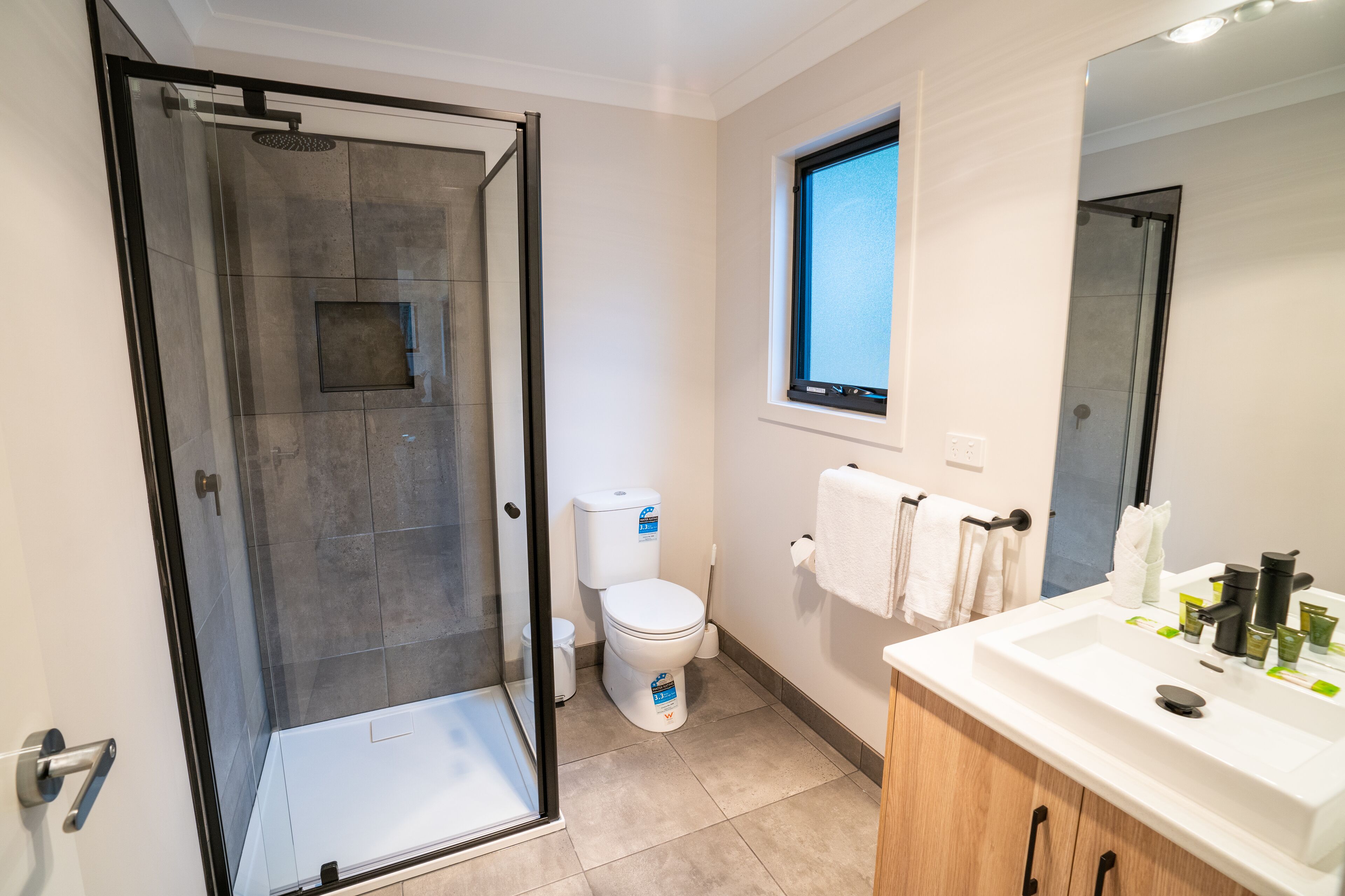 Une salle de bain moderne avec une douche à parois de verre, des toilettes, un lavabo avec des articles de toilette et une fenêtre.