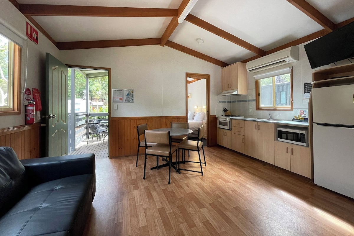 A compact kitchen with wooden cabinets, a dining area with a table and chairs, and a black leather sofa.