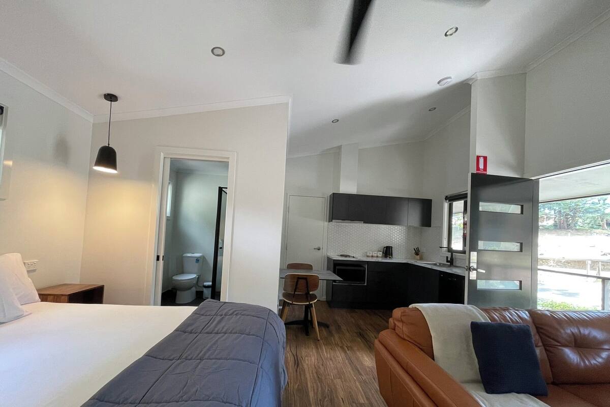 A modern hotel room with a large bed, a sofa, a small kitchen area, and a bathroom.