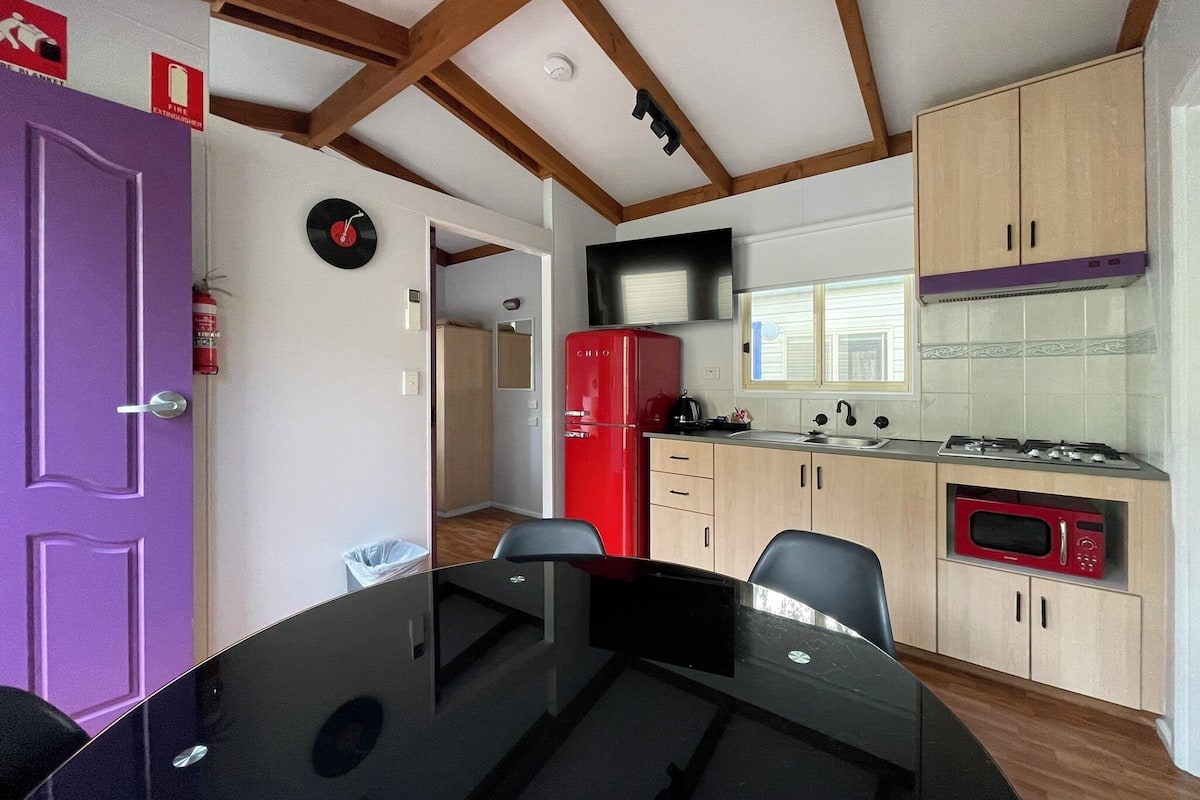 A compact kitchen with a dining table, wooden cabinets, a red refrigerator, and a microwave.