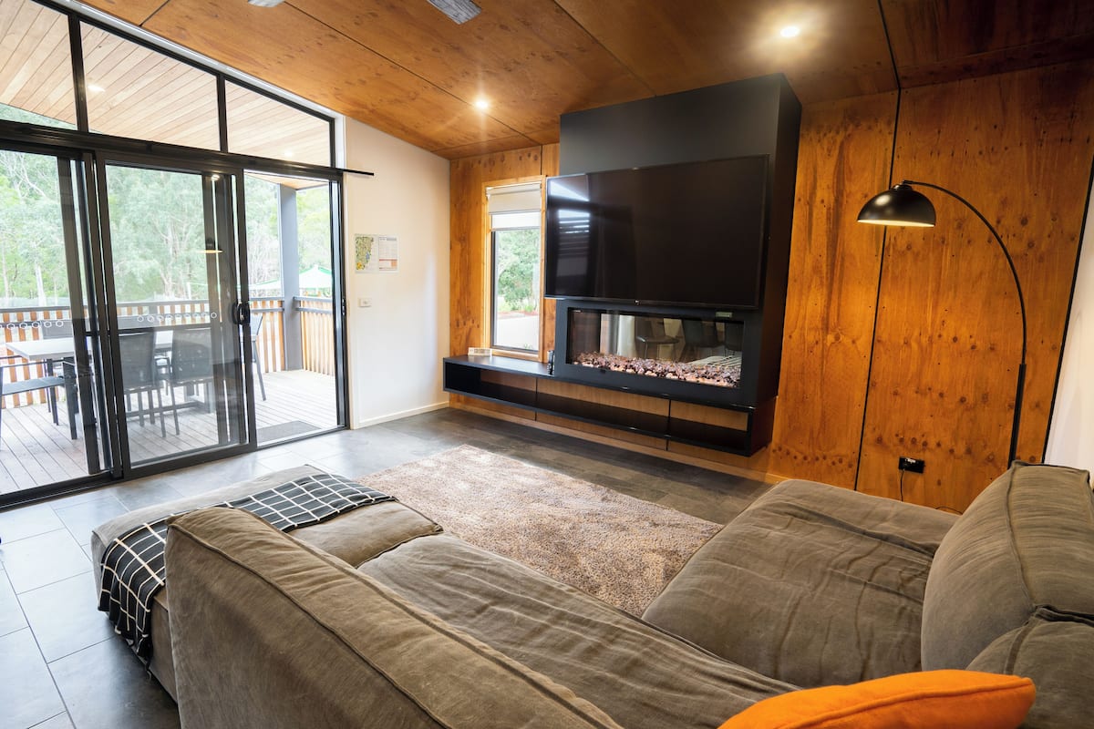 A modern living room with a large flat-screen TV, a fireplace, and a sliding glass door leading to an outdoor deck.