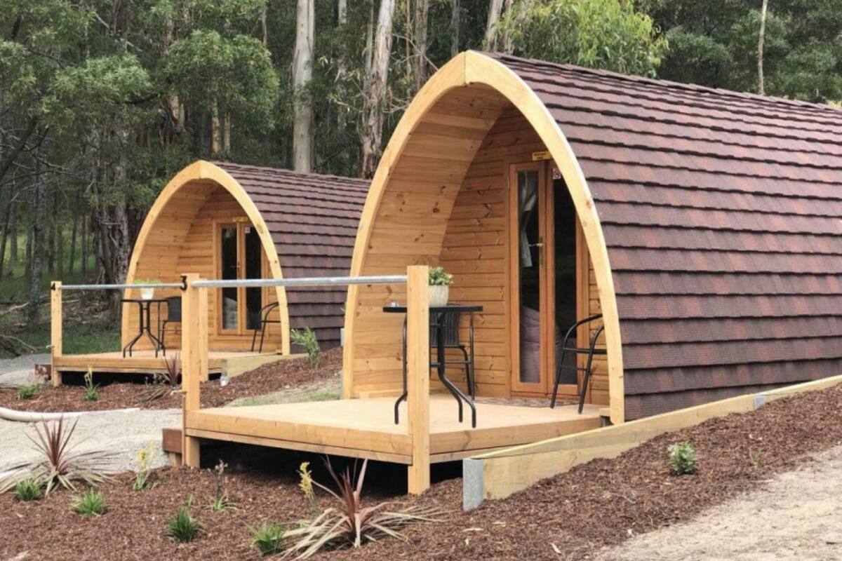 Two wooden cabins with arched doorways, each featuring a small deck with a table and chairs.