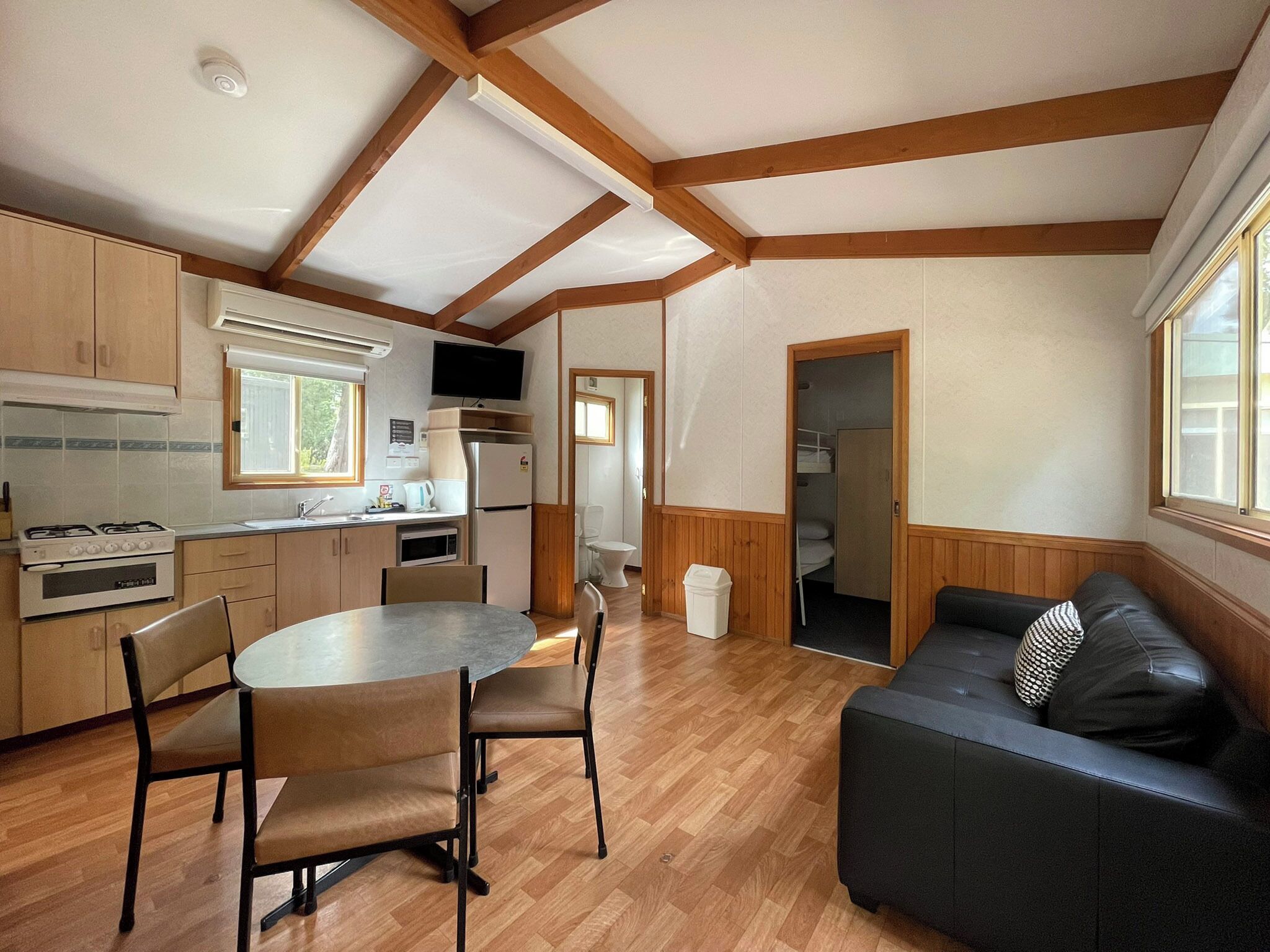 Una cocina compacta con vigas de madera, una mesa de comedor con sillas, un sofá de cuero negro y un baño pequeño visible a través de una puerta abierta.